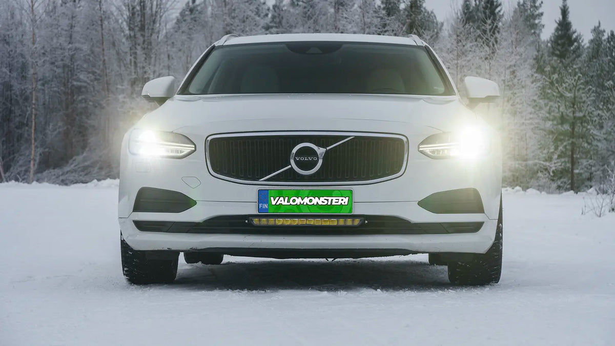 Valkoinen henkilöauto lumisessa maisemassa, jonka puskuriin on asennettu palava led-lisävalopaneeli.